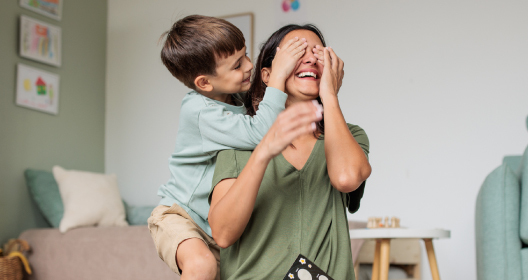 A boy stands behind his smiling mother and playfully covers her eyes with his hands.